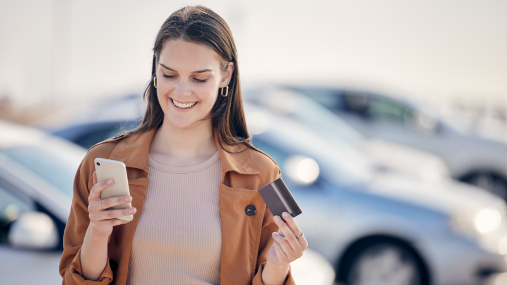 Junge Frau mit Smartphone vor einem Mietwagen